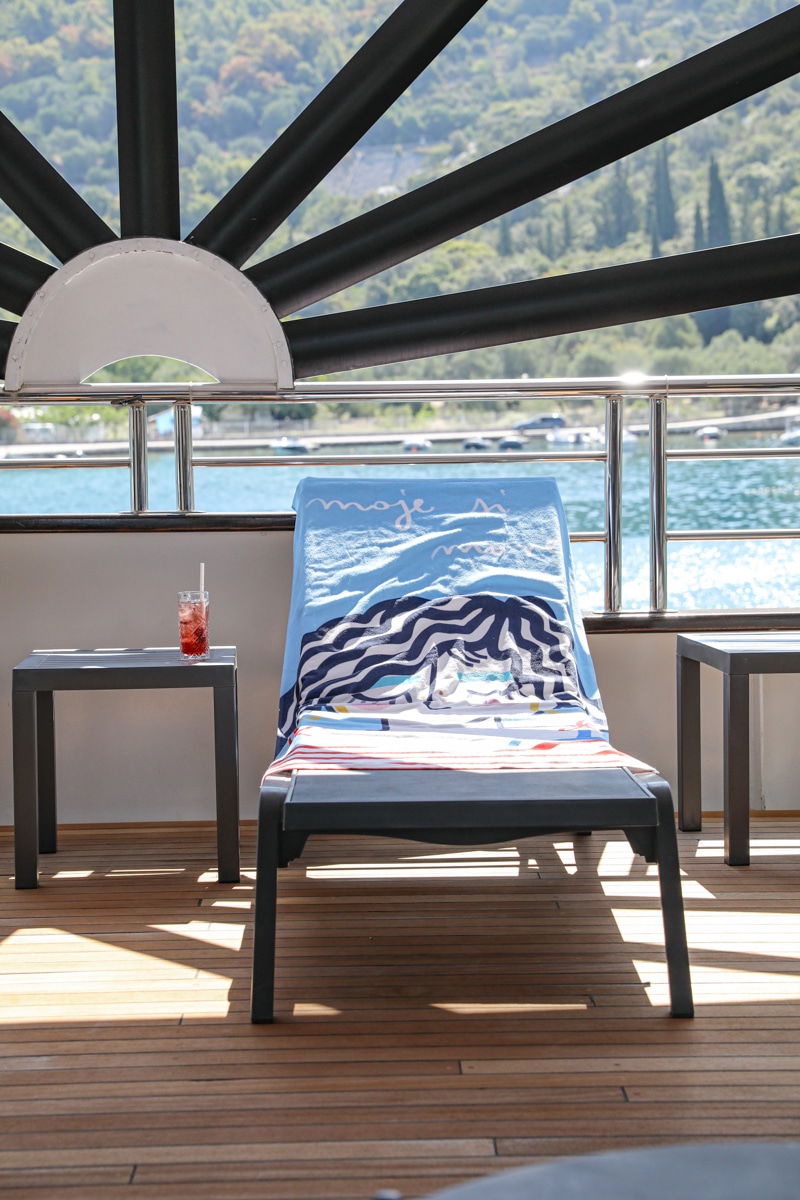 A sun lounger with a printed towel is positioned on a deck with tables holding drinks. The background reveals a scenic view of the Adriatic Sea and rolling hills, inviting guests to unwind after swimming.