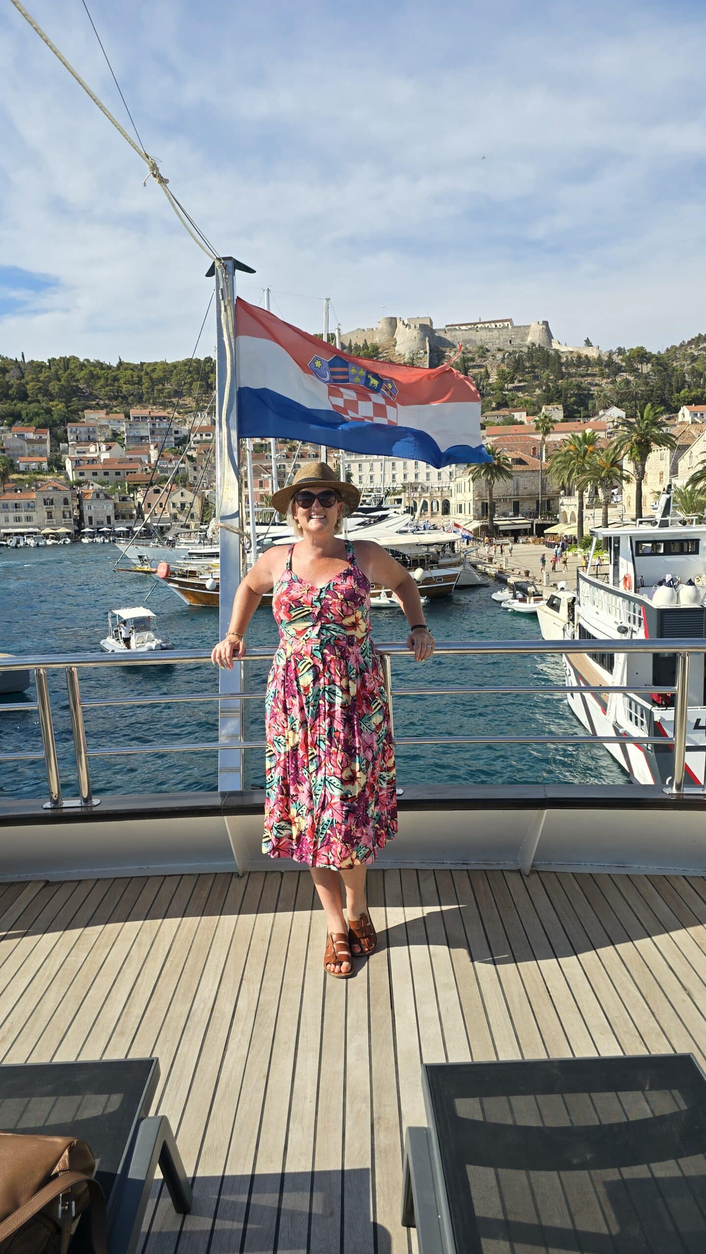 SJ in a pink floral dress and straw hat stands on the deck of a boat, with a Croatian flag behind her. In the background is Hvar Island