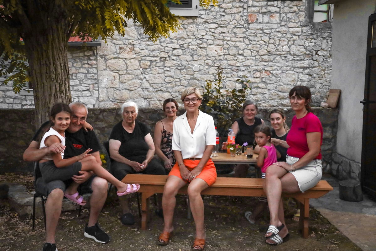 A group of seven people sits outdoors on wooden benches in front of a stone building in Drnis - prsut tasting experience